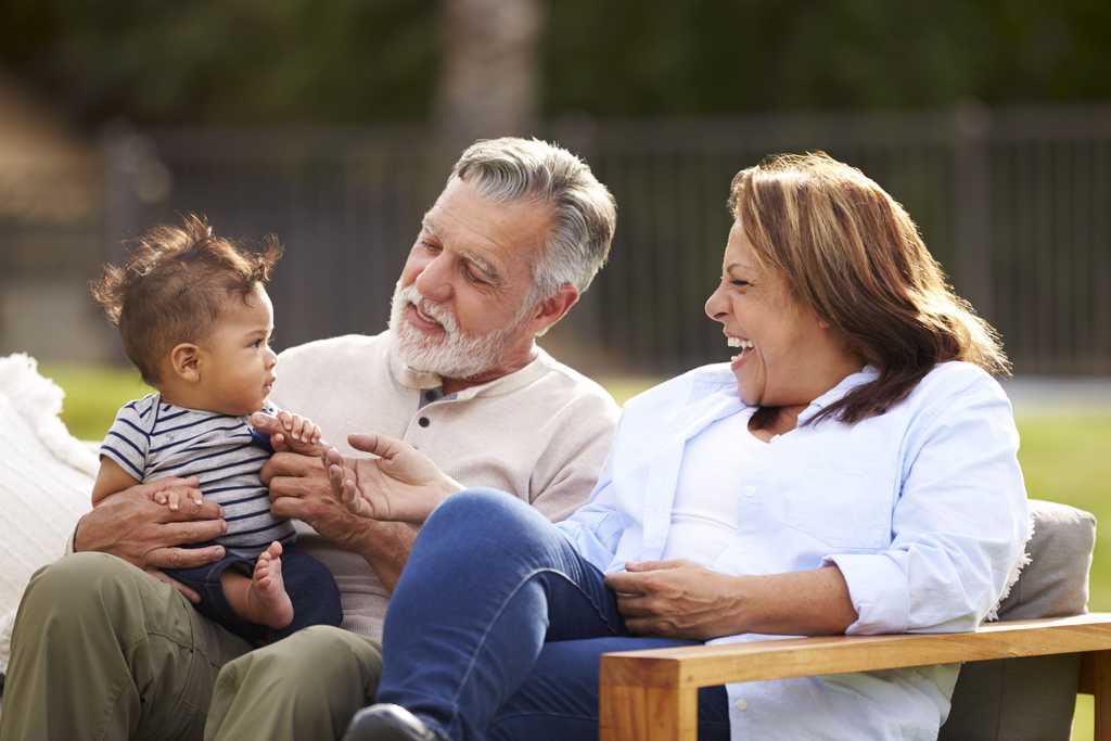 Senior couple sitting in the garden with their baby grandson, smiling at him