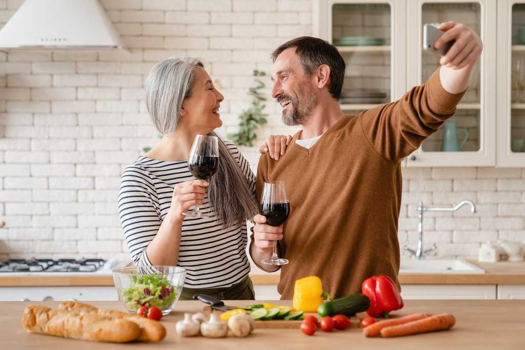 A couple in their 50s in their kitchen, each holding up a glass of wine and look romantically into each other's faces while taking a selfie with a smart phone
