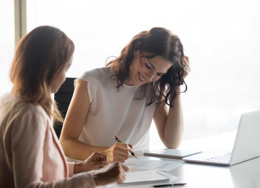 two professional women in their 40s look over paperwork with an open laptop in an office setting.