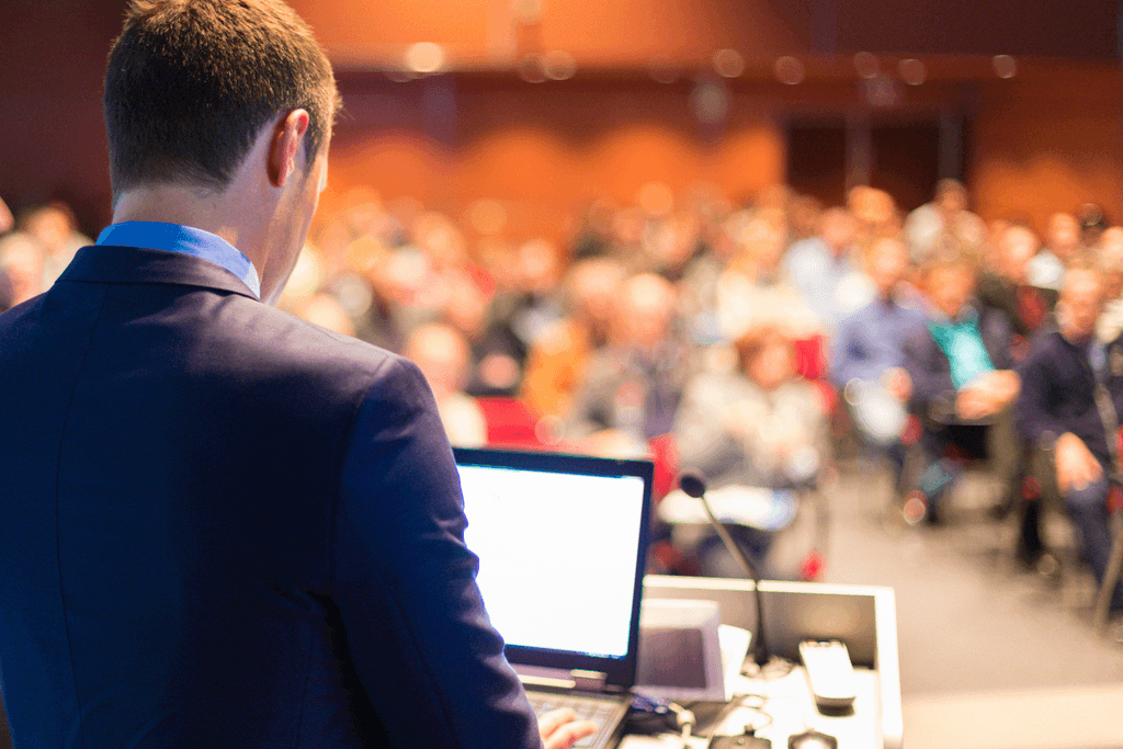 Back of man standing at a podium with an open laptop, looking out into a medium size audience in an auditorium.