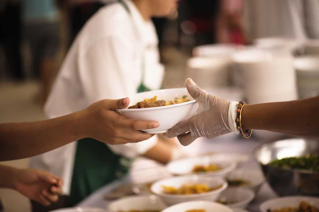A person handing a bowl of food to another person in a soup kitchen type setting, showing only their hands.